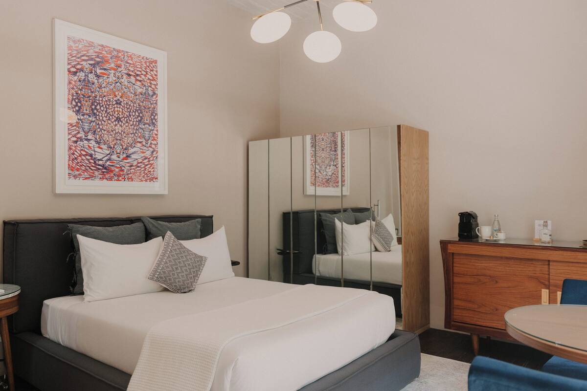 A modern hotel room with a large bed, a wooden dresser, a round dining table, and a large abstract painting on the wall.