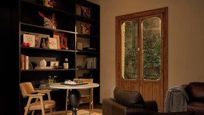 Een gezellige woonkamer met een houten boekenkast, een ronde tafel met schaakstukken en een groot raam met uitzicht op groen.