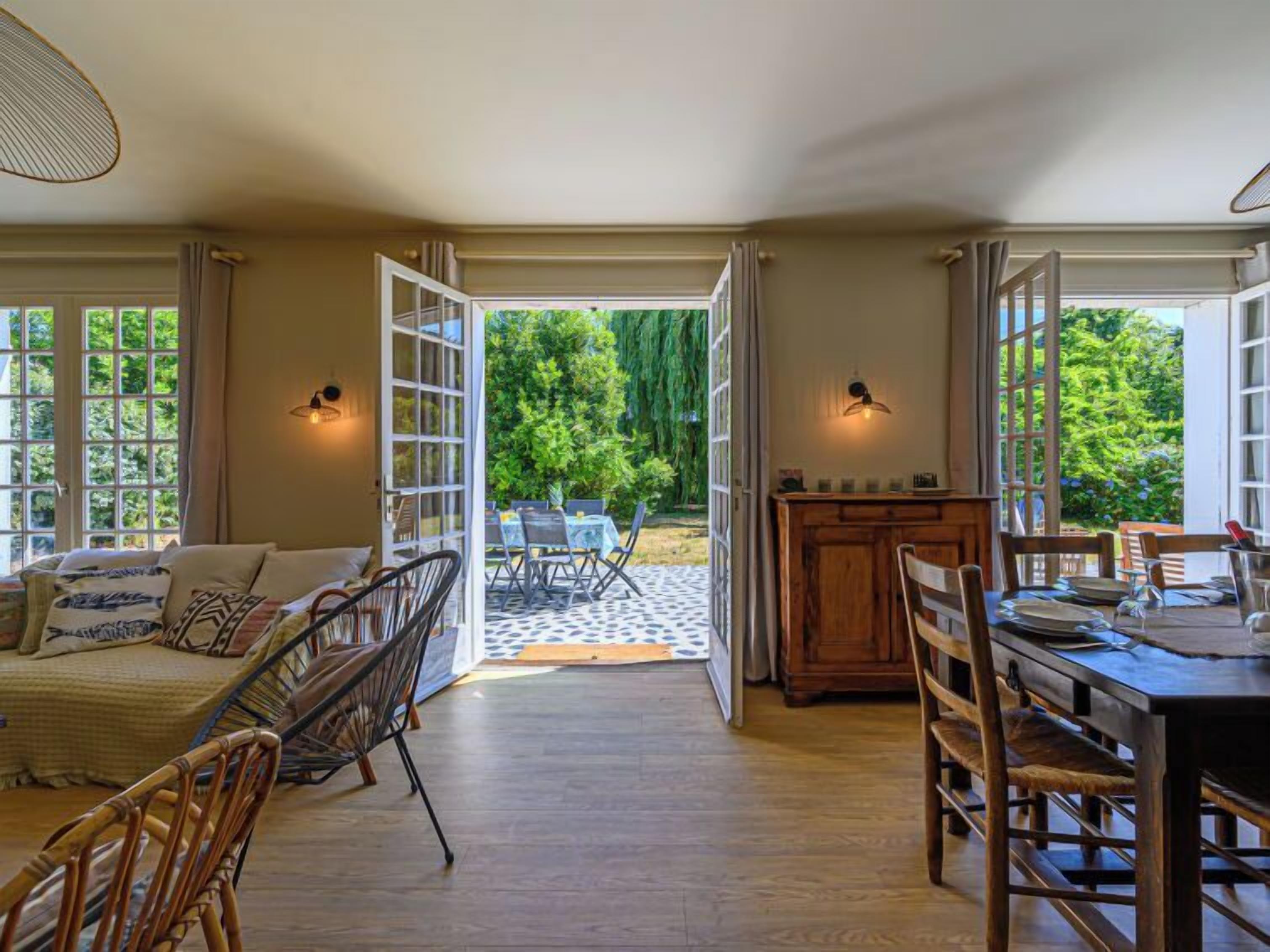 An open-plan living area with a dining table and chairs, a sofa, and a view of a garden through large windows.