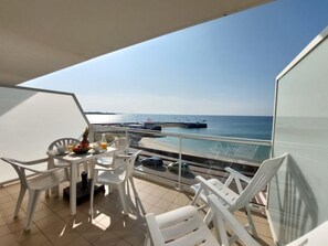 Uma varanda com uma mesa posta para o café da manhã, cadeiras de plástico branco e vista para o mar e barcos.