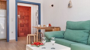 Una sala de estar con un sofá verde, una mesa blanca con un frutero y copas de vino, un taburete de madera y una puerta de madera con marco azul.