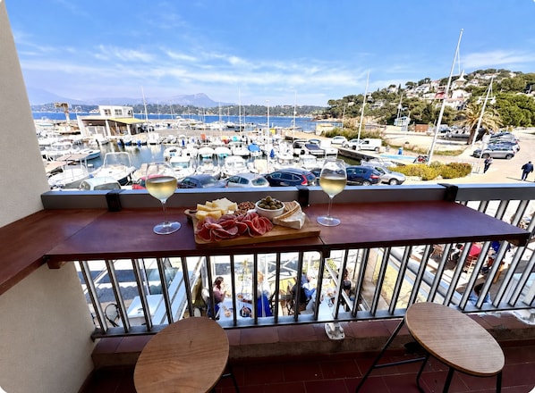 A balcony with a view of a marina, a wooden table with a cheese board and wine glasses, and two chairs.