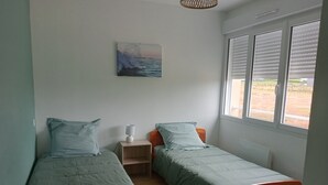 Una habitación con dos camas individuales, una mesita de noche con lámpara, un cuadro con una vista marina y una ventana con persianas.