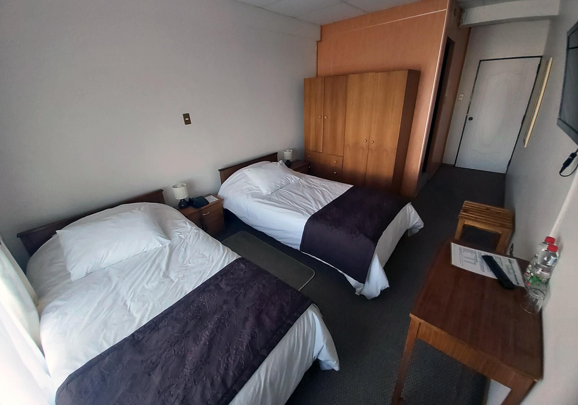 A hotel room with two beds, a wooden wardrobe, a small table with a bottle, and a door.
