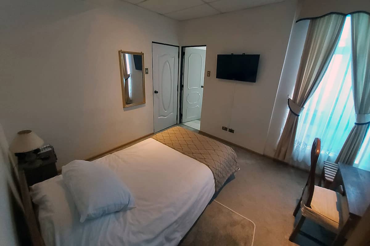 Habitación de hotel con cama, escritorio con lámpara, televisor en la pared y ventana con cortinas.