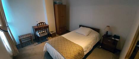 A bedroom with a bed, a nightstand, a desk with a chair, a wardrobe, and a small trash bin.