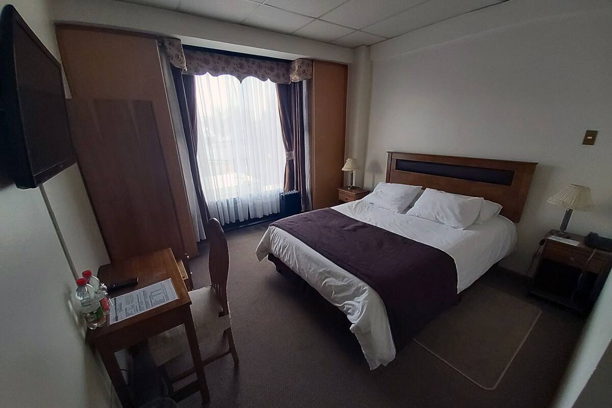 Habitación de hotel con cama, un escritorio con silla, televisor montado en la pared y ventana con cortinas.
