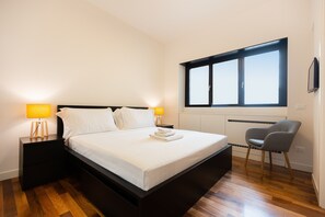Cama bem arrumada com lençóis brancos, duas luminárias de cabeceira, uma cadeira e uma janela grande com persianas.