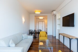 Uma sala de estar moderna com um sofá, uma poltrona amarela, uma mesa de centro de vidro e uma televisão na parede.