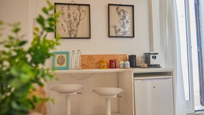 A compact kitchen with a refrigerator, a coffee machine, and a small countertop with various items. There are two white bar stools and a potted plant in the foreground. - Circe Studio Sea View (Syracuse)