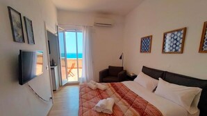 A hotel room with a bed, a TV, a chair, and a view of the beach through the door. - Circe Studio Sea View (Syracuse)