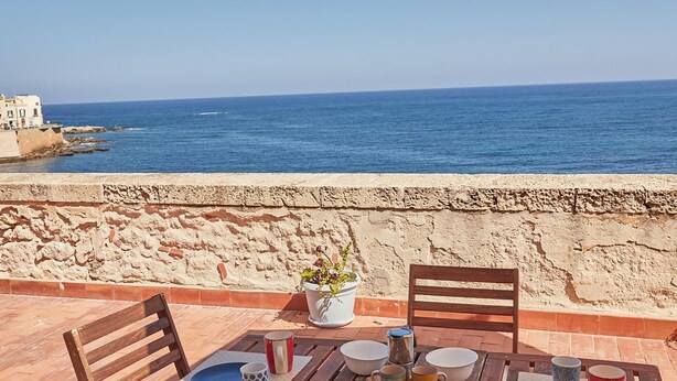 A wooden table with chairs and a beige umbrella, overlooking a coastal view with a stone wall and buildings in the background. - Circe Studio Sea View (Syracuse)