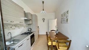 A compact kitchen with a gas stove, sink, and modern cabinetry. A dining table with chairs is positioned in the center of the room. - Casa La Palma in San Giuliano Terme (Vecchiano)