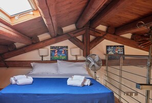 A bedroom with a sloped ceiling, wooden beams, a bed with a blue bedspread, white pillows, two rolled towels, a wall-mounted fan, and two paintings. - Cala Rossa in Siracusa (Syracuse)