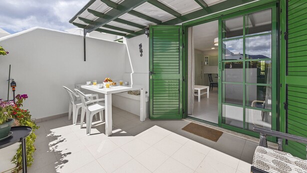Une terrasse avec une table et des chaises blanches, une porte verte et une vue sur l’intérieur.