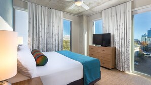 A hotel room with a bed, a TV on a dresser, and large windows with curtains.