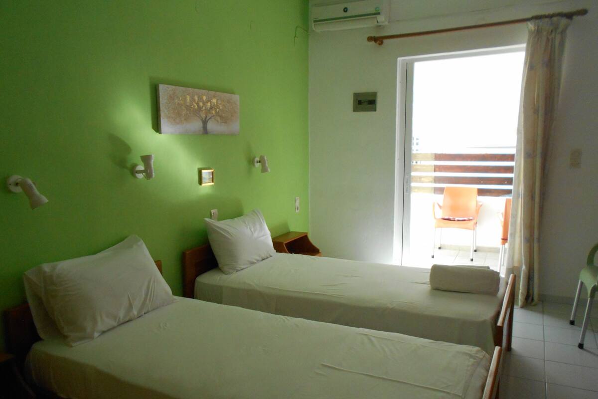 A room with two beds, a green wall, a window with curtains, and a small table with a chair.