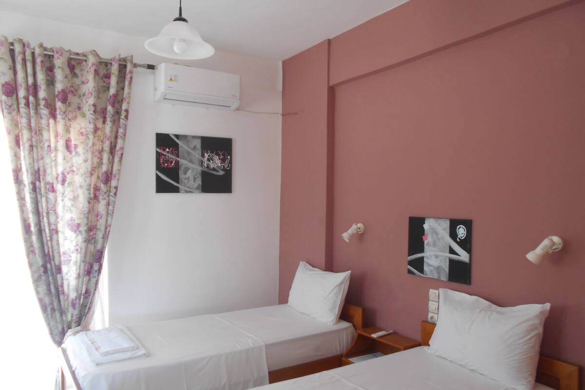 A room with two single beds, a window with floral curtains, an air conditioning unit, and two wall-mounted light fixtures.