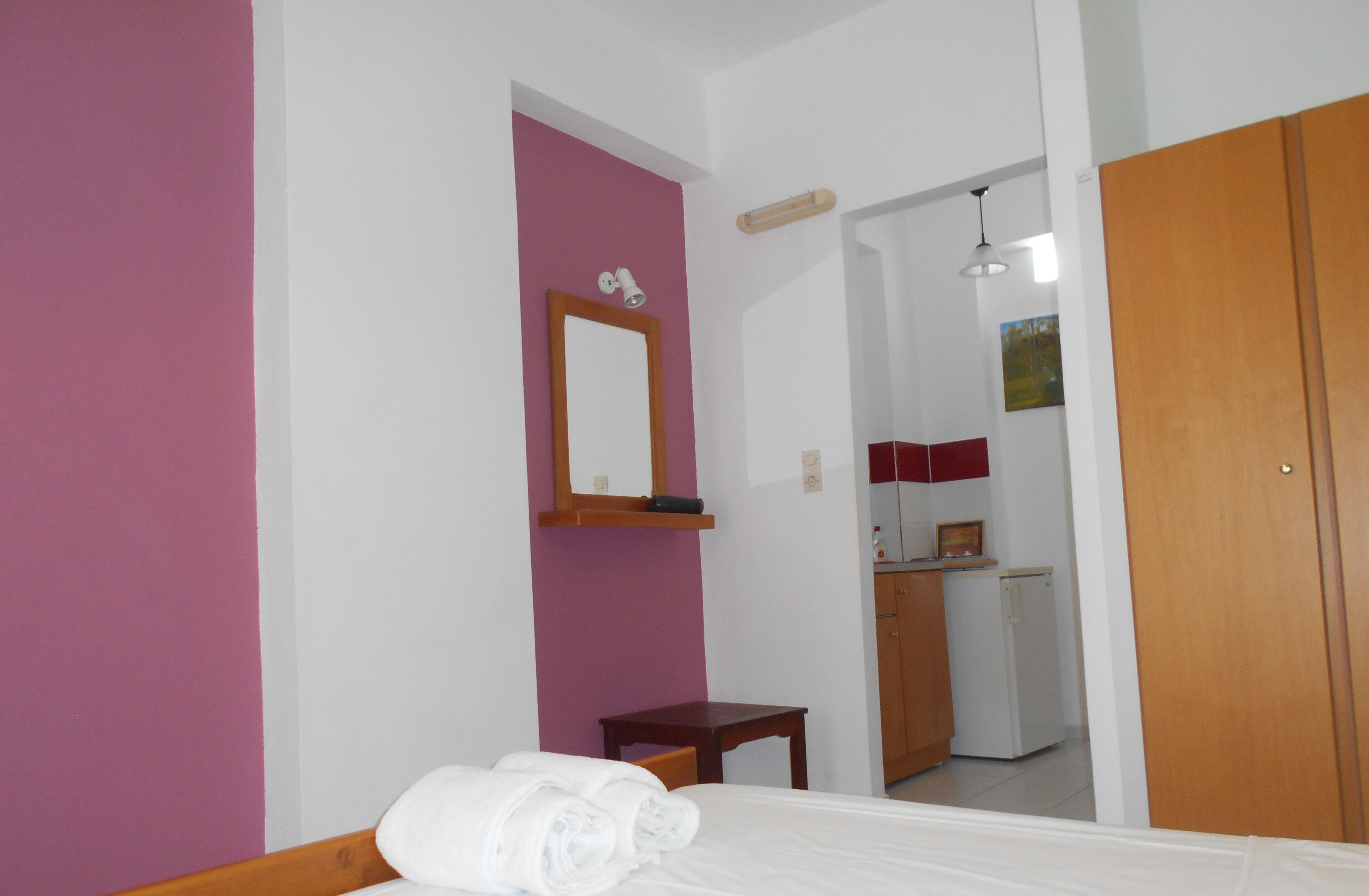 Une chambre d’hôtel avec un lit, une petite table, un miroir, une porte et, au fond, une kitchenette.