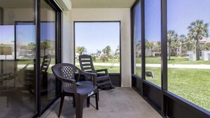 A balcony with two chairs and a table, large windows, and a view of palm trees and a lawn. - Ocean Village Club-Sleeps 4 with First  Floor Access and Screen Porch (St. Augustine)