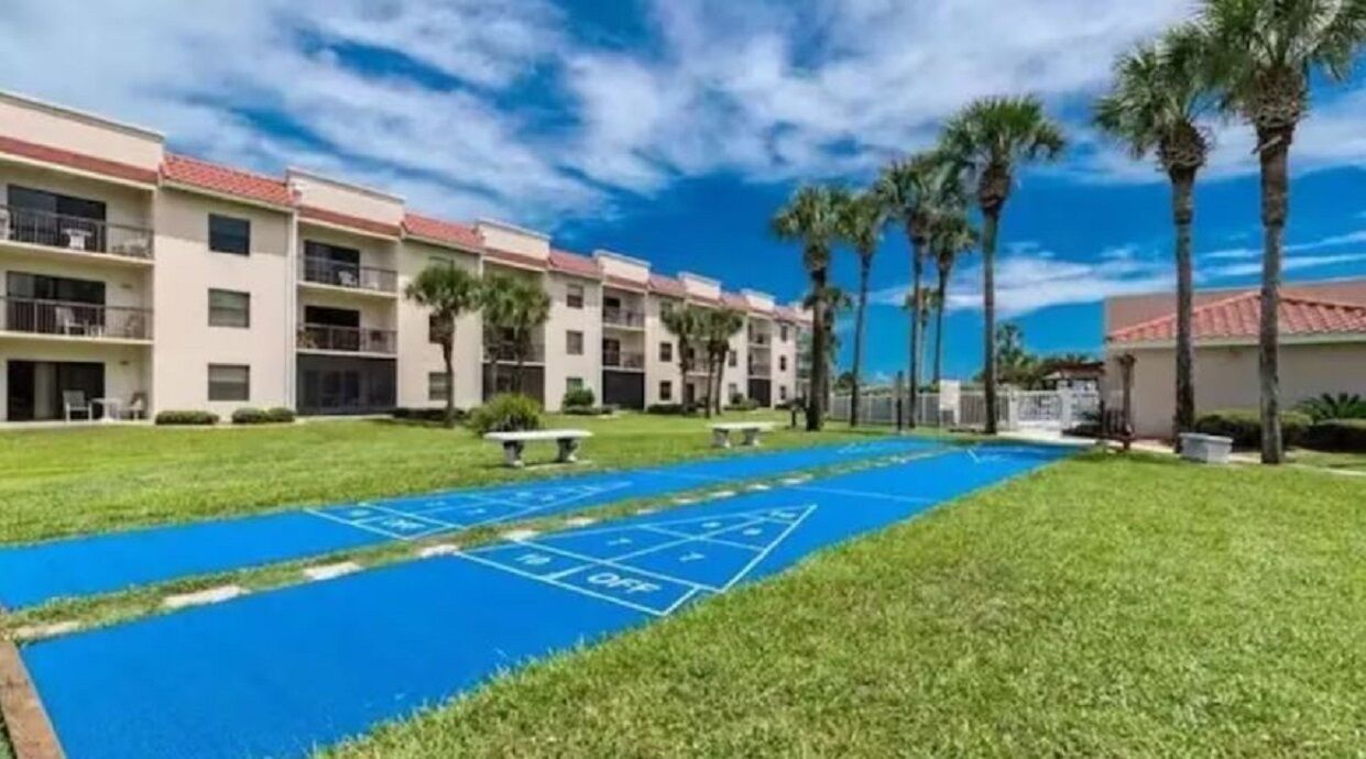 A courtyard with a shuffleboard court, palm trees, and a row of multi-story buildings with balconies.