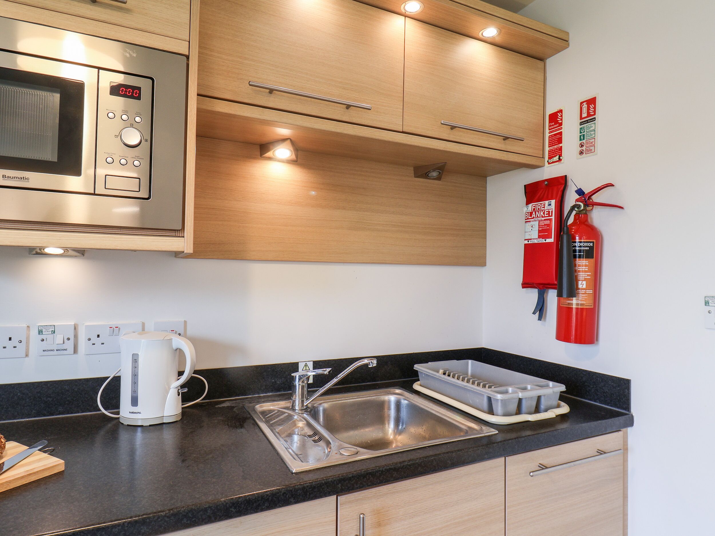 A kitchen with a microwave, sink, and cabinets. There are fire extinguishers on the wall.
