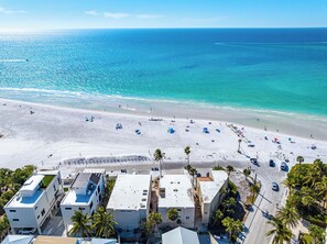 An aerial view of a beach with white sand, clear blue water, and a few buildings in the foreground.