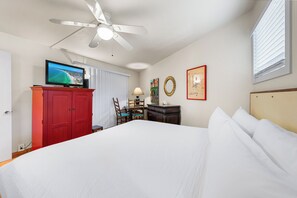 Une chambre d’hôtel avec un lit, un téléviseur posé sur une commode rouge, un ventilateur de plafond, un bureau avec une chaise et des œuvres d’art accrochées aux murs.