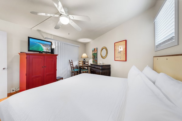 A hotel room with a bed, a television on a red cabinet, a ceiling fan, a desk with a chair, and artwork on the walls.