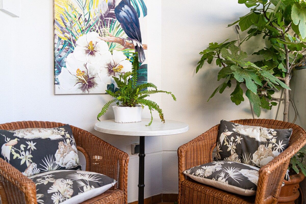 A corner room with wicker chairs, a small round table, a potted plant, and a large floral painting on the wall.
