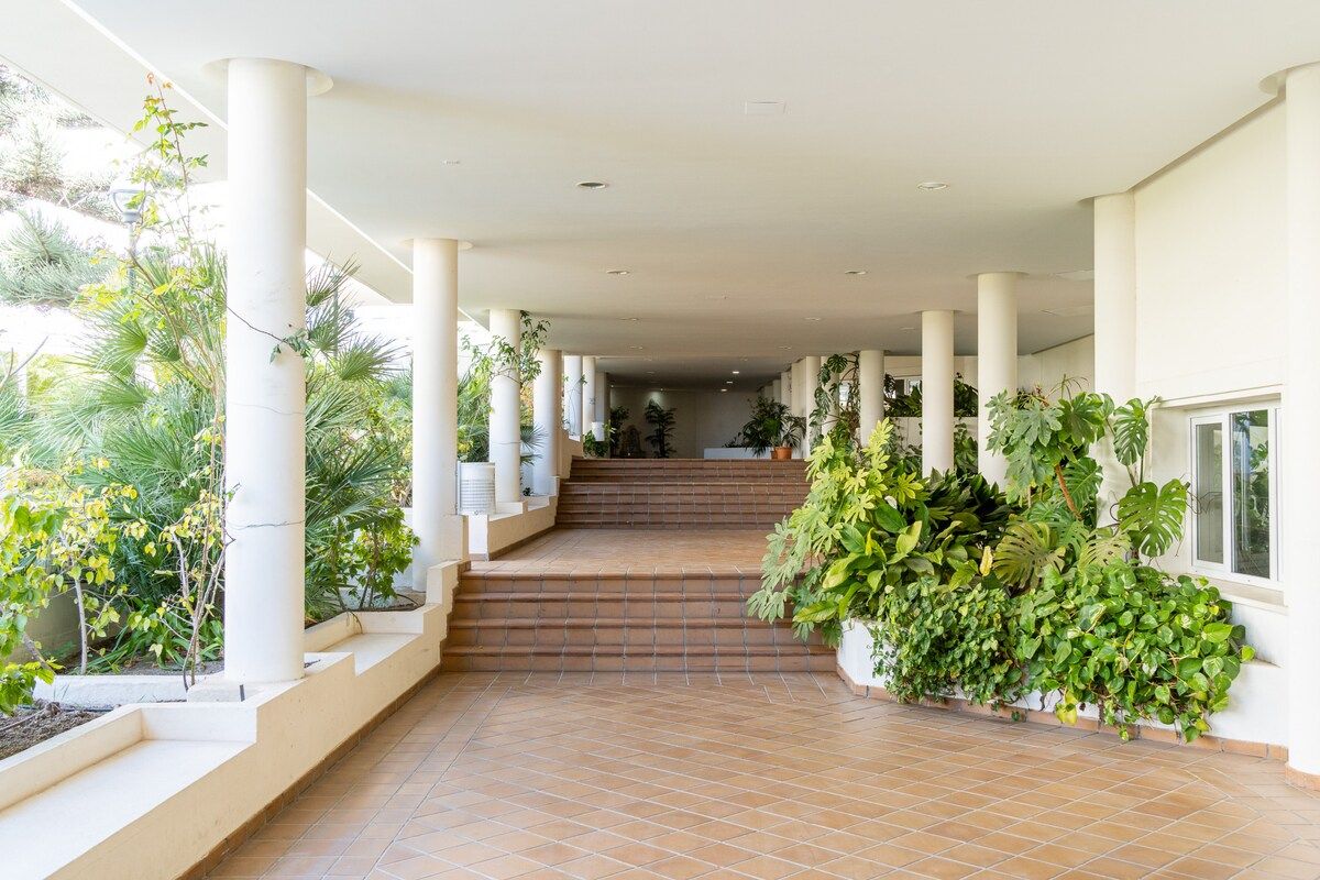 A covered walkway with steps leading to a higher level, flanked by columns and potted plants.