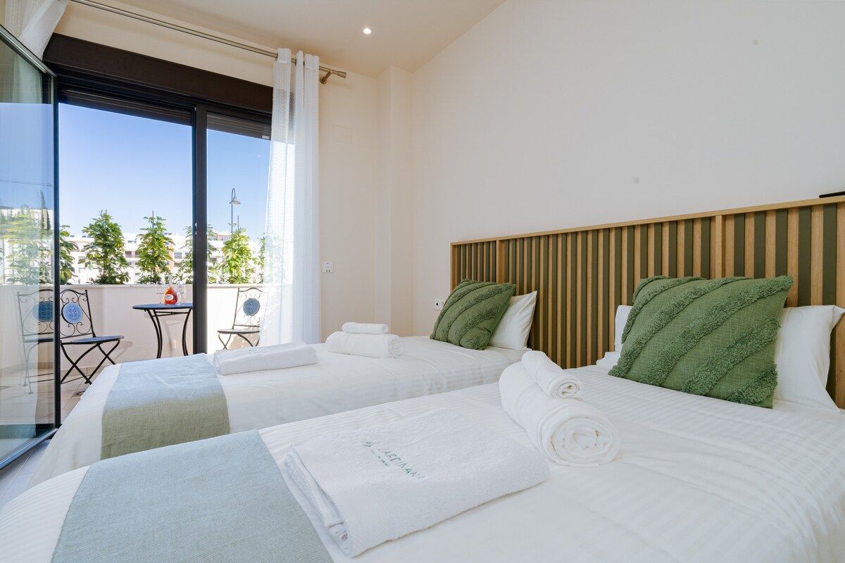 Habitación de hotel con dos camas, vista a la ciudad y balcón con mobiliario exterior.
