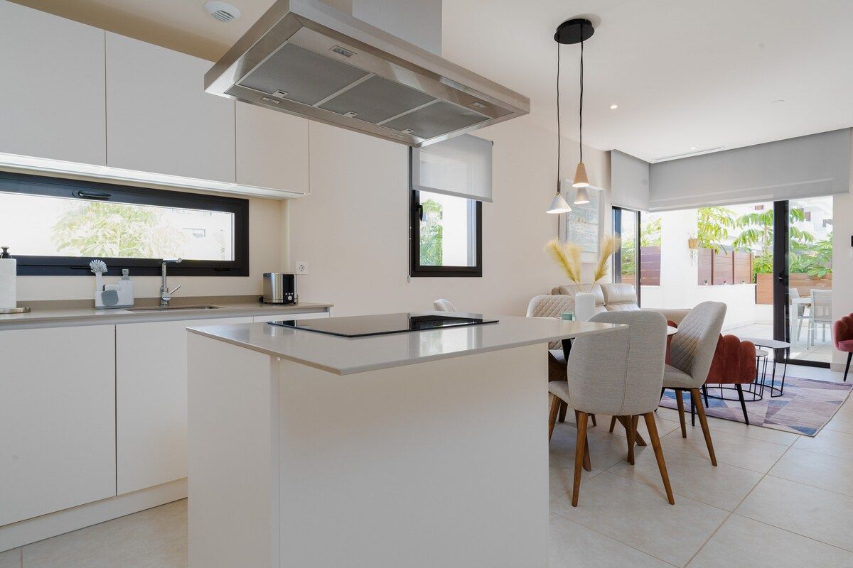 A modern kitchen with a central island, white cabinetry, and a dining area with chairs and a table.