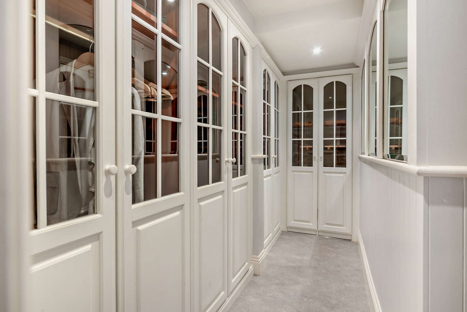 Un couloir étroit aux murs et aux portes recouverts de panneaux blancs, qui mène à un espace de rangement avec une tringle pour suspendre les vêtements.