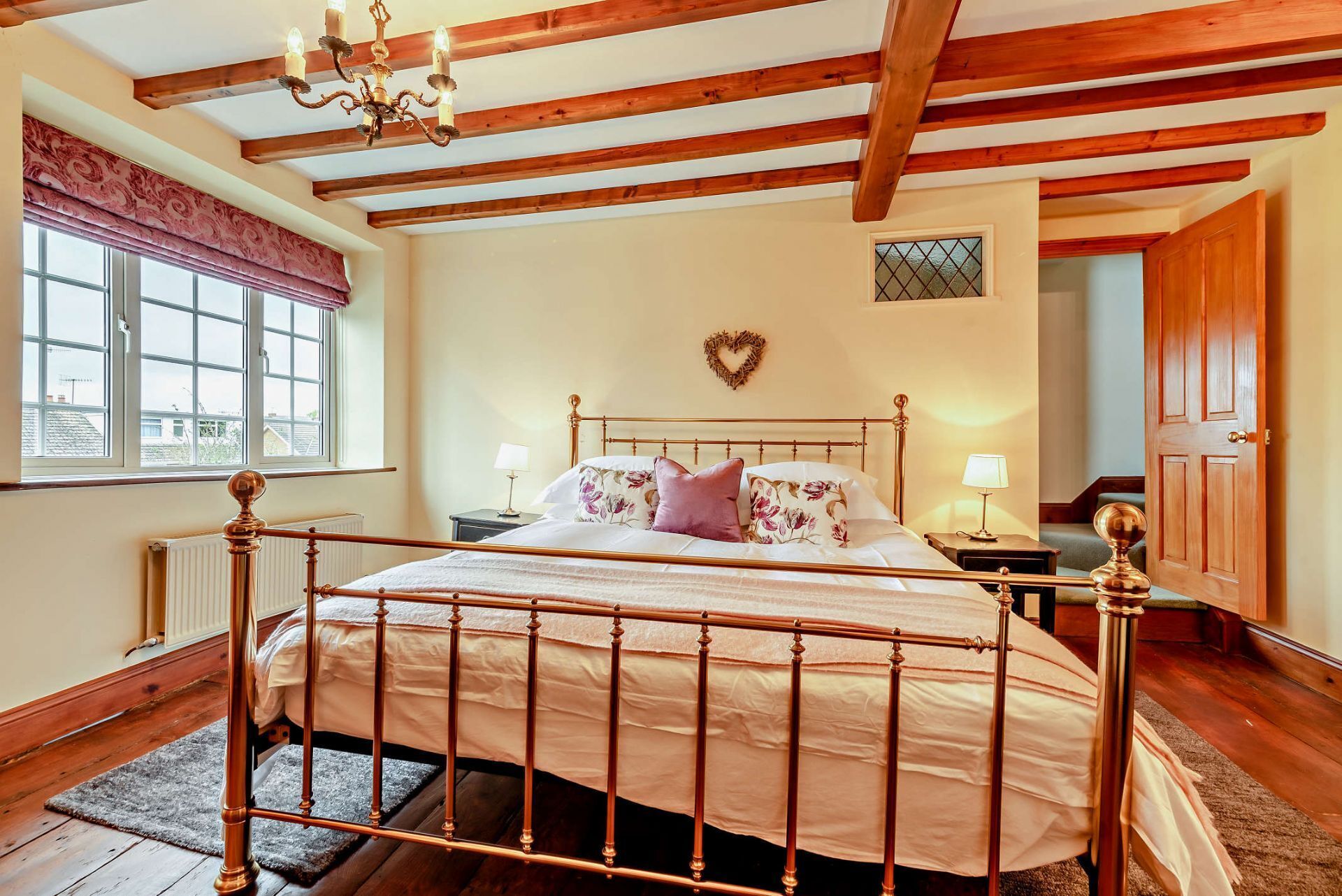Un dormitorio con estructura de cama de madera, una araña y una decoración con forma de corazón en la pared, además de una ventana con cortinas.