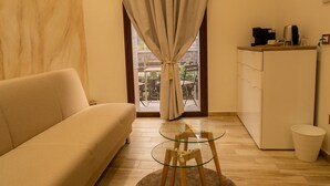Un salon comprenant un canapé, une table en verre avec des pieds en bois, un tapis, une fenêtre avec des rideaux et un coin cuisine équipé d’appareils électroménagers.
