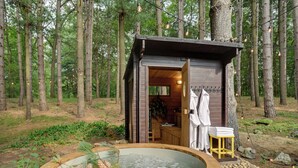 A rustic cabin with a hot tub, wooden deck, and a small outdoor shower area surrounded by trees. - Magical Metamora Cottage w/ Fire Pit, Pond, Deck! (Metamora)