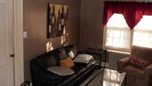 A living room with a black leather sofa, a glass coffee table, a beige armchair, a red curtain, and a painting of wine bottles on the wall. - Windsor Lofts 2 (Roanoke)