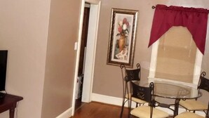 A dining area with a round glass table and four chairs, a framed picture on the wall, and a window with a red valance. - Windsor Lofts 2 (Roanoke)