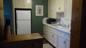 Une cuisine de taille réduite avec des meubles blancs, un réfrigérateur blanc, une table en bois et un évier équipé d’un égouttoir à vaisselle.