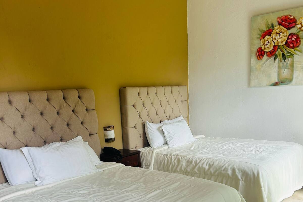 Habitación de hotel con dos camas, una pared amarilla y un cuadro de flores.