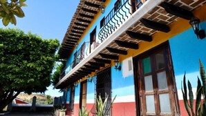Un edificio con fachada azul y amarilla, un balcón con barandillas negras y una ventana con puerta de madera.