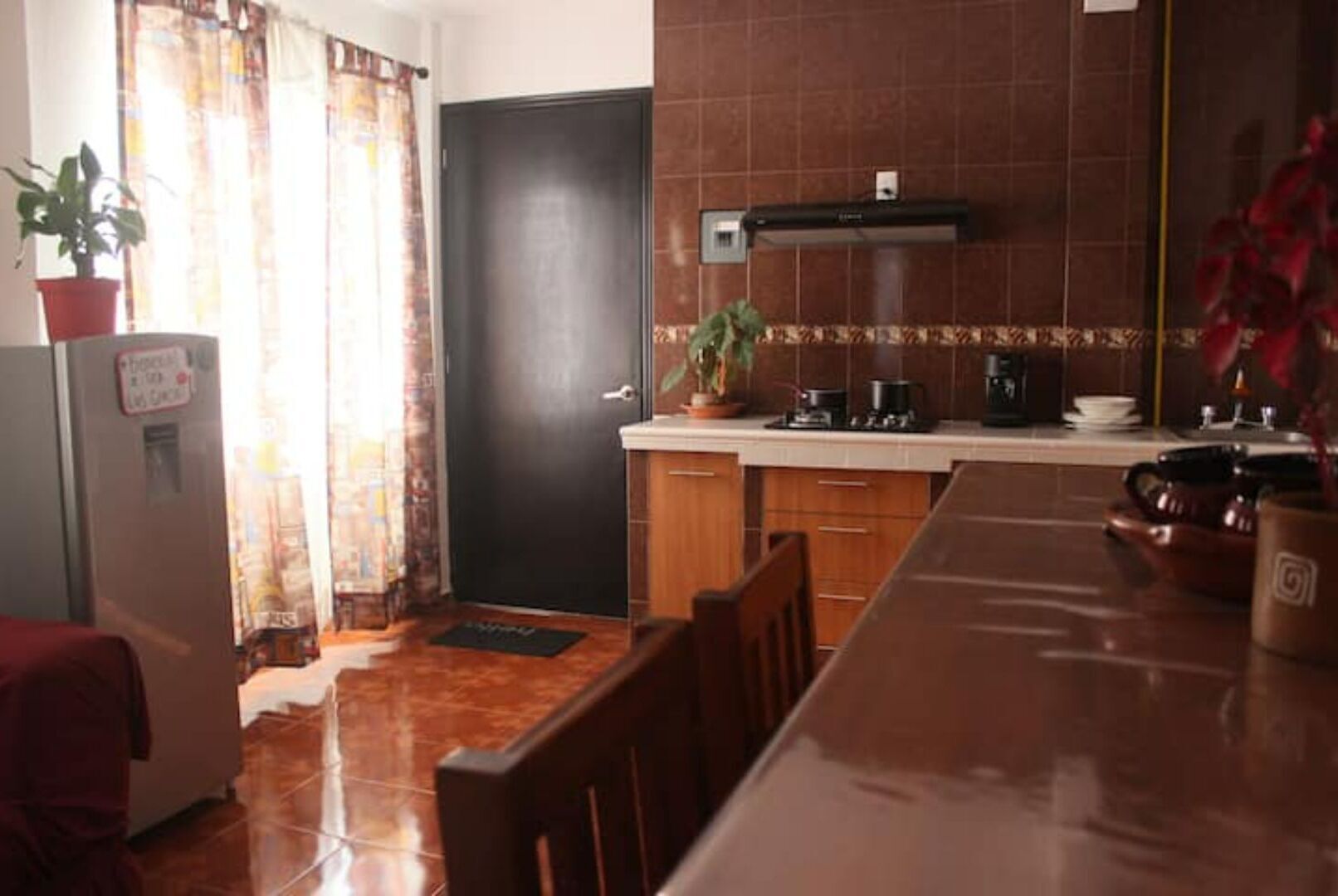 A compact kitchen with a refrigerator, stove, and sink. There is a dining table with chairs and a potted plant on the counter.