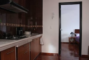 Una cucina con mobili scuri in legno, un fornello e un lavandino. Si apre una porta verso una camera da letto con un letto e un comodino.