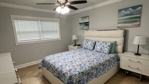 Une chambre à coucher avec un lit, des tables de chevet, un ventilateur de plafond, une fenêtre avec des stores et un tableau au mur.