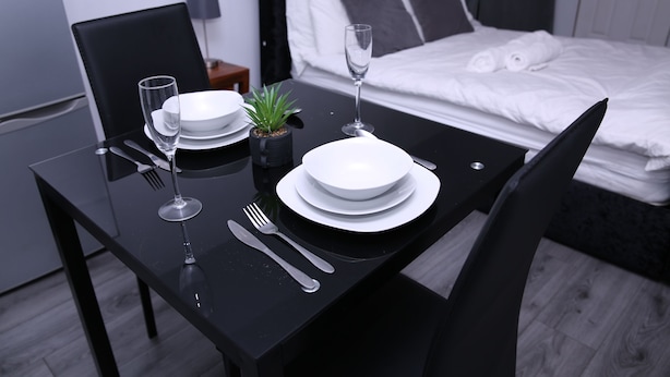 A neatly set dining table with plates, cutlery, and a wine glass in a modern room with a bed in the background. - The Lodge Oldham (Oldham)