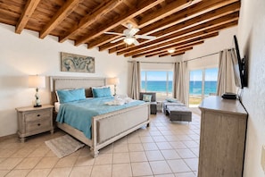 Une chambre à coucher avec un lit, des tables de chevet, un ventilateur de plafond, un tableau, une vue sur la mer et un téléviseur.