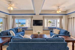 A spacious living room with two sofas facing a flat-screen TV, two ceiling fans, and large windows with curtains.