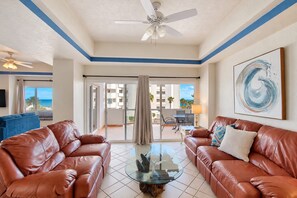 A hotel room with a balcony, leather sofas, a glass coffee table, a ceiling fan, and a painting.