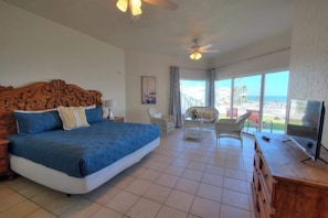A bedroom with a large bed, a ceiling fan, a TV, and a view of the beach.